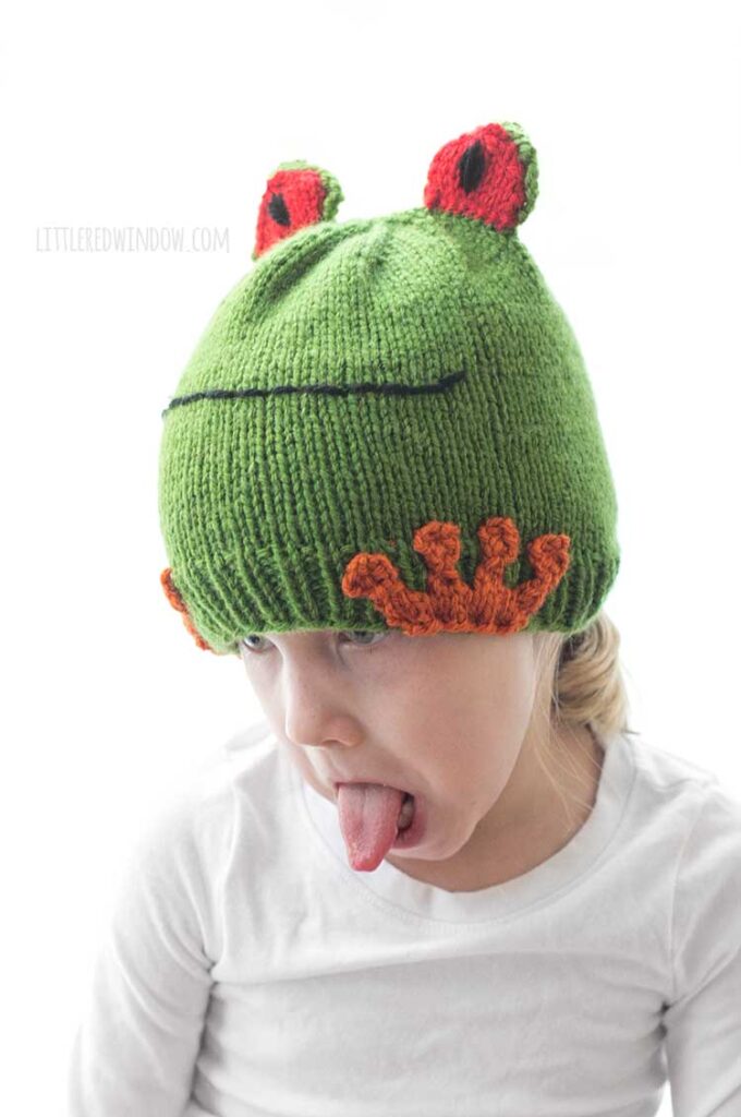little girl wearing knit tree frog hat and looking down with her tongue sticking out like a frog