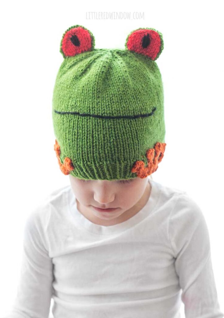 little girl wearing a white shirt and colorful tree frog hat and looking down at her lap