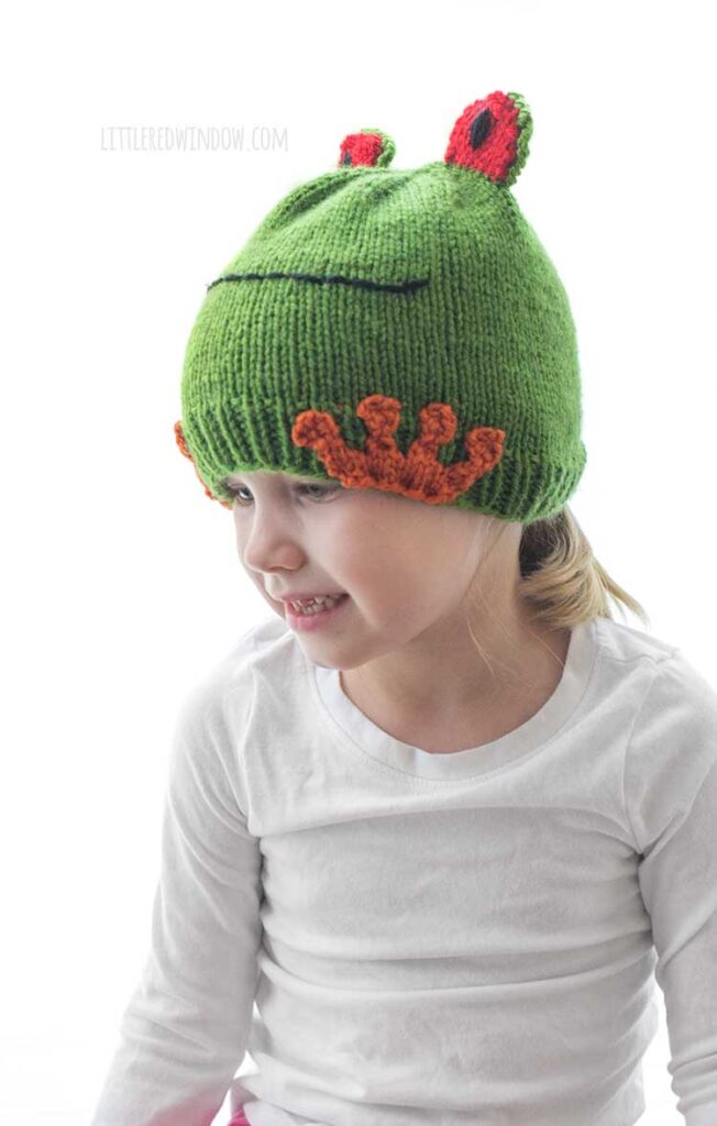 little girl wearing a white shirt and colorful tree frog hat and looking off to the left