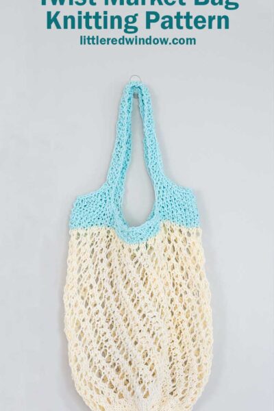 off white lace knit market bag with a twisting stitch pattern and light blue handles hanging in front of a gray wall