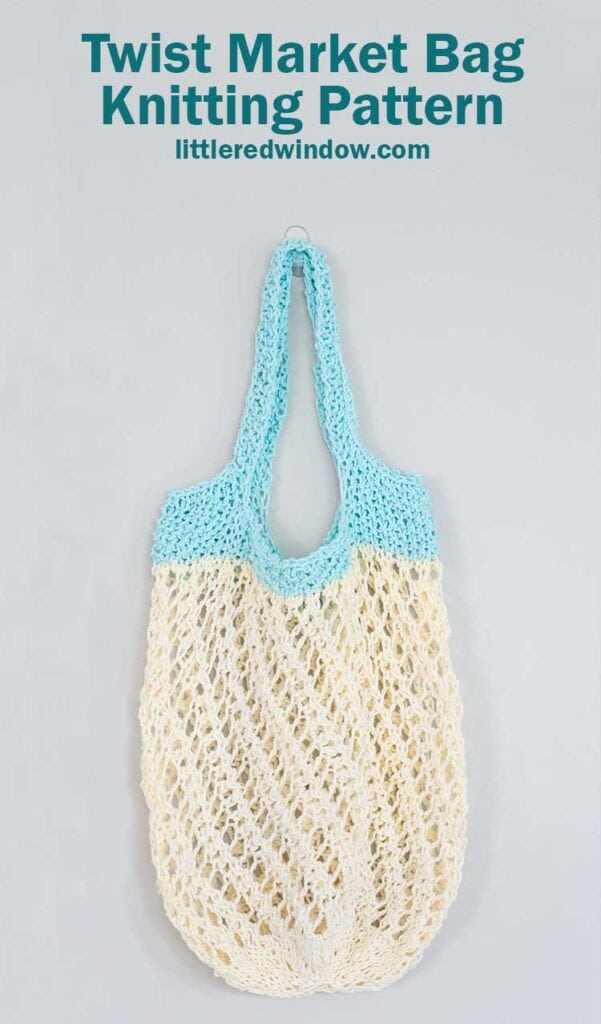off white lace knit market bag with a twisting stitch pattern and light blue handles hanging in front of a gray wall