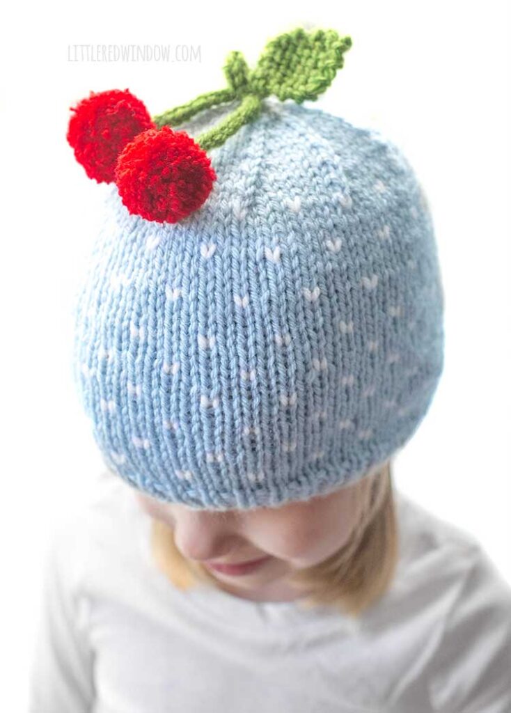 closeup of two red cherries stem and leaf on top of a light blue and white polka dot knit hat