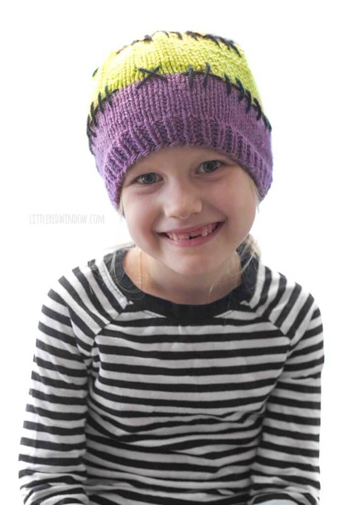 child in black and white striped shirt wearing a purple acid green and orange colorblocked knit hat with black stitching lines between the colors looking straight ahead and smiling in front of a white background