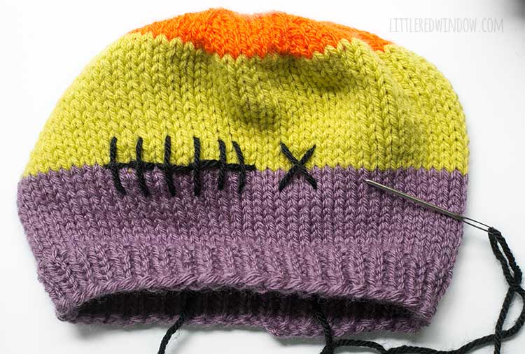 progress shot showing finished Dr Frankenstein style black yarn stitches between the purple and green stripes on a knit baby hat in front of a white background