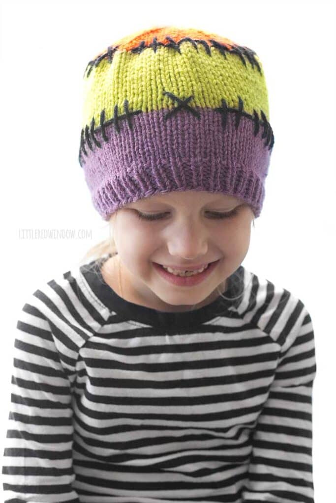 child in black and white striped shirt wearing a purple acid green and orange colorblocked knit hat with black stitching lines between the colors looking down and smiling in front of a white background