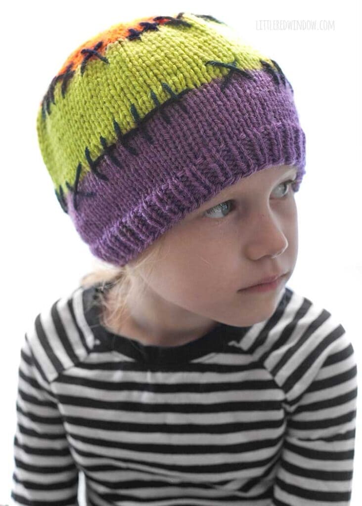 child in black and white striped shirt wearing a purple acid green and orange colorblocked knit hat with black stitching lines between the colors looking off in the distance to the right in front of a white background