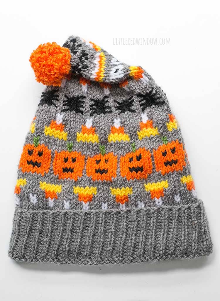 flat lay image of gray knit halloween stocking cap with pumpkin candy corn and spiders patterns on it with the top folded downlaying on a white background