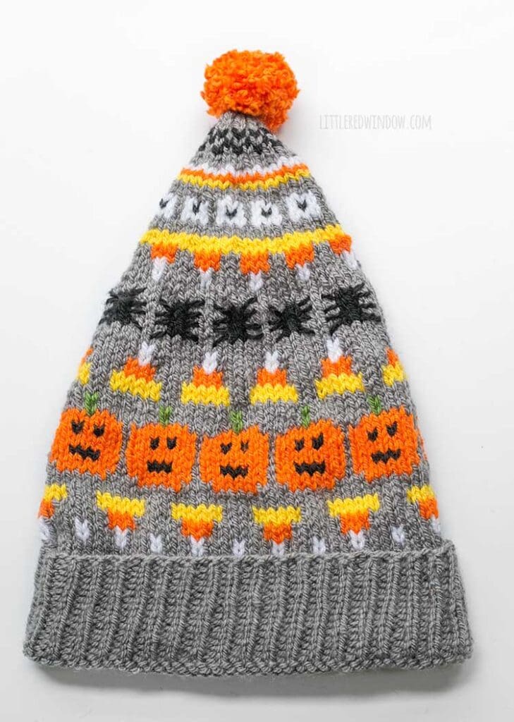 flat lay image of gray knit halloween stocking cap with pumpkin candy corn and spiders patterns on it laying on a white background