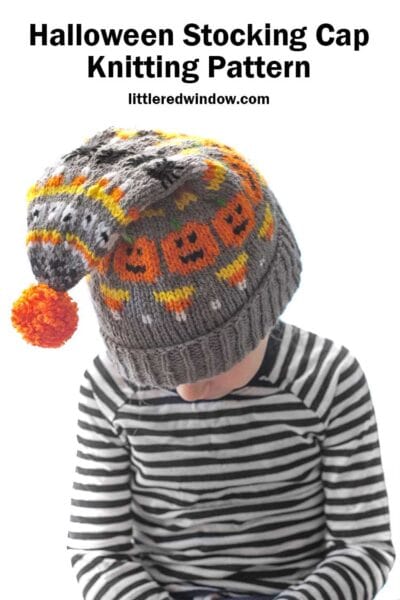 little girl in black and white striped shirt with gray knit stocking cap with halloween items knit on it in orange yellow and black and with an orange pom pom flopping forward and she leans down