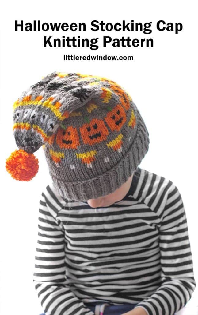 little girl in black and white striped shirt with gray knit stocking cap with halloween items knit on it in orange yellow and black and with an orange pom pom flopping forward and she leans down