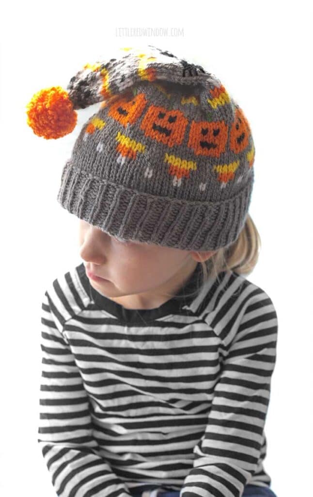 little girl in gray stocking cap knit with candy corns and jack o lanterns and spiders and an orange pom pon on top look off to the left