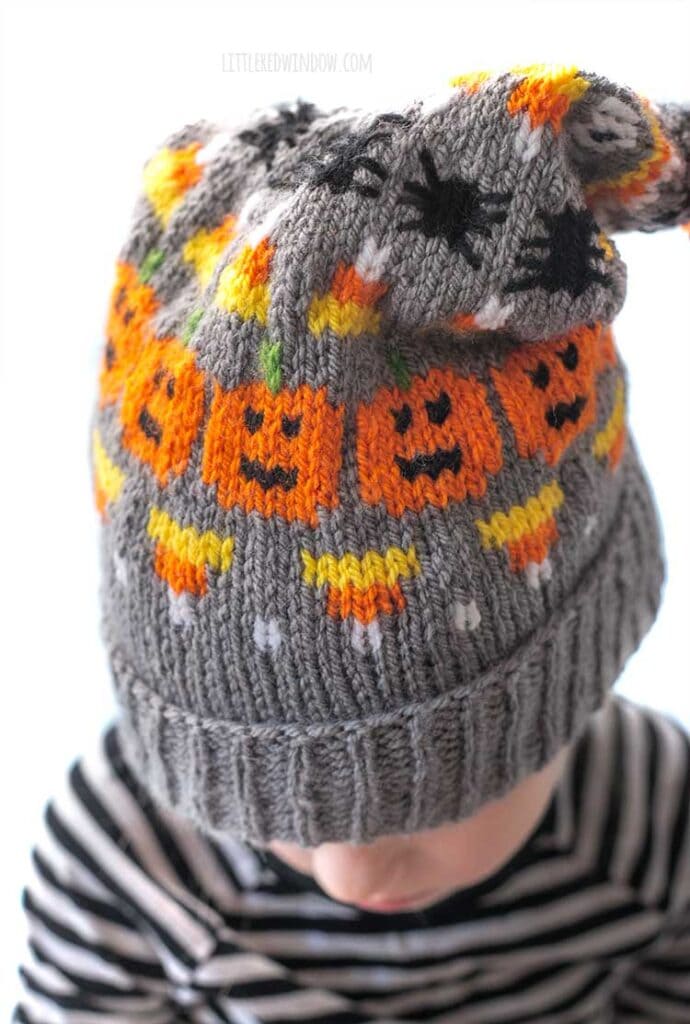 closeup of little girl in gray stocking cap knit with candy corns and jack o lanterns and spiders and an orange pom pon on top look down with the pom pom in back
