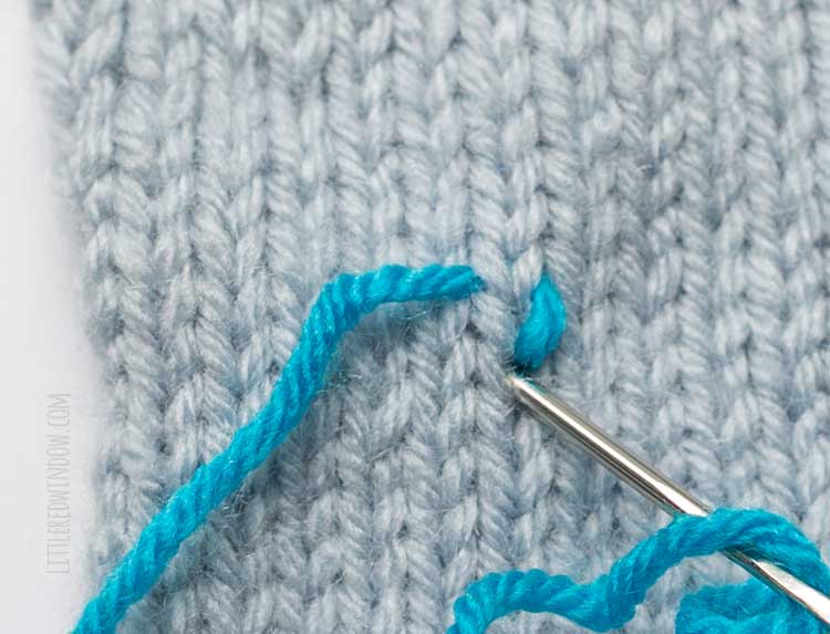 showing where to insert the yarn needle to complete one duplicate stitch on light blue stockinette stitch swatch