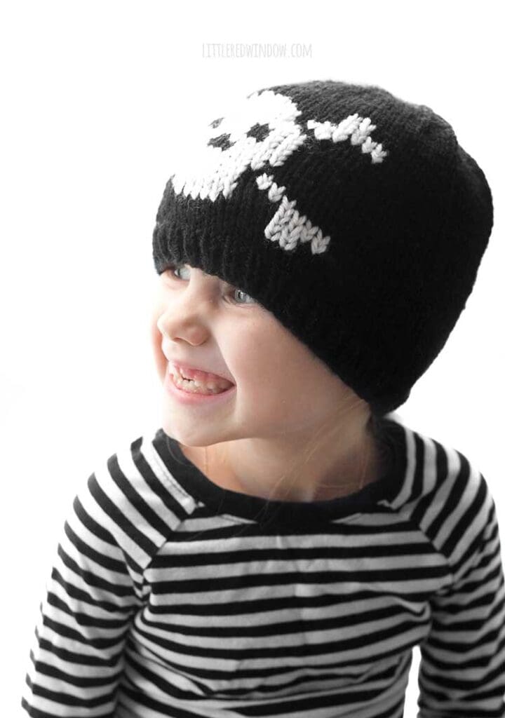 child in black and white striped shirt wearing a black knit hat with a white skull and cross bones knit on the front and looking off to the left and smiling