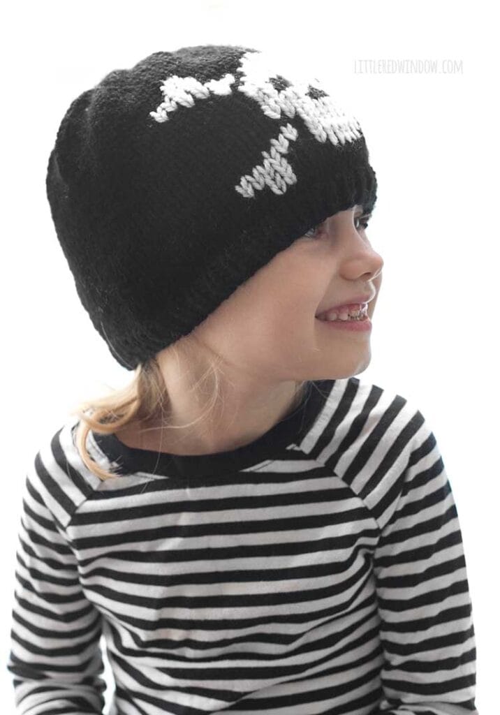 child in black and white striped shirt wearing a black knit hat with a white skull and cross bones knit on the front and looking off to the right and smiling