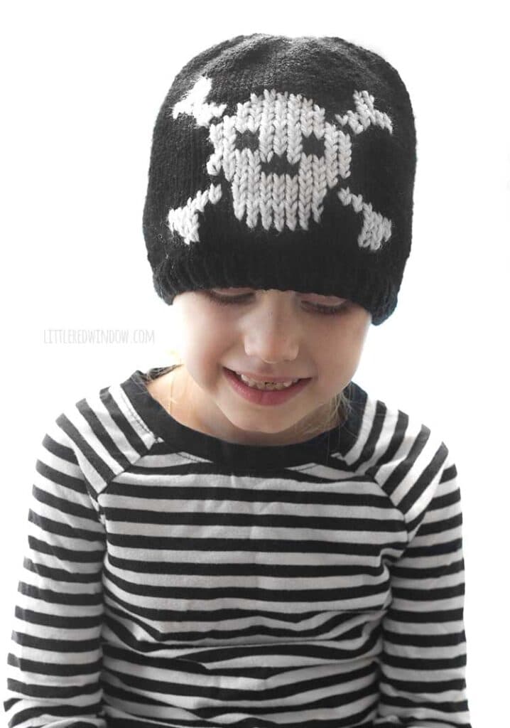 child in black and white striped shirt wearing a black knit hat with a white skull and cross bones knit on the front and looking down nd smiling