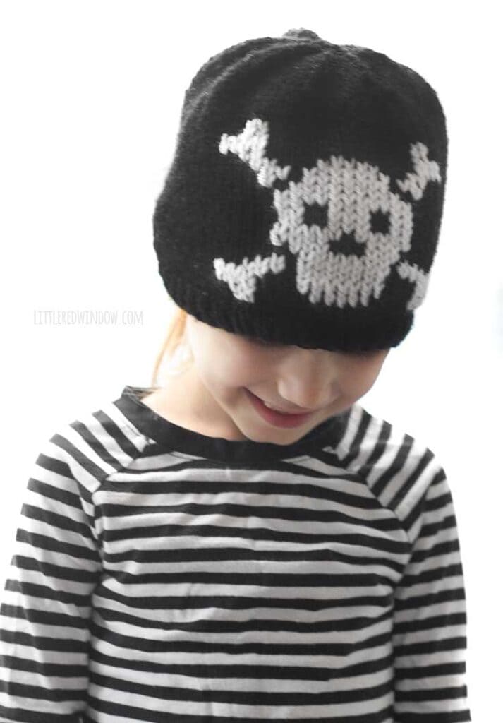 child in black and white striped shirt wearing a black knit hat with a white skull and cross bones knit on the front and looking down and right and smiling