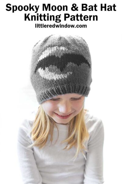 little girl in white shirt wearing gray knit hat with white crescent moon and black bat looking down at her lap and smiling