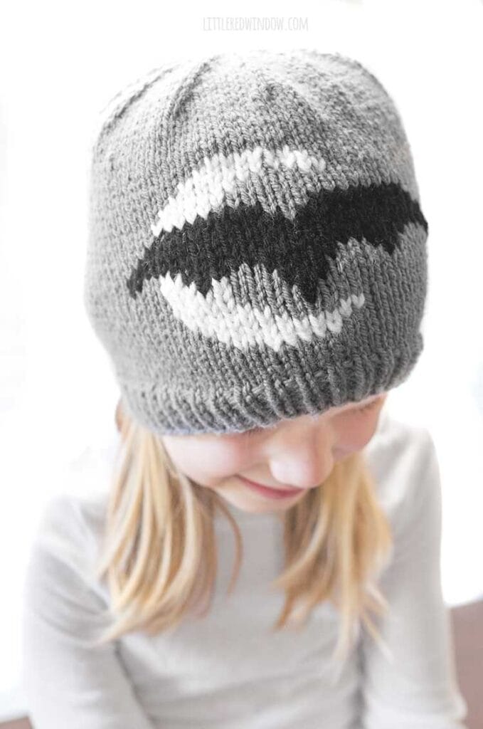 closeup of little girl in white shirt wearing gray knit hat with white crescent moon and black bat looking down at her lap and smiling