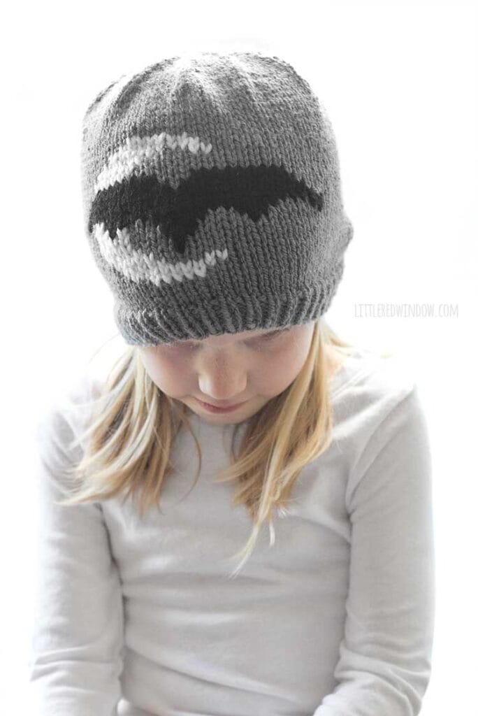little girl in white shirt wearing gray knit hat with white crescent moon and black bat looking down at her lap