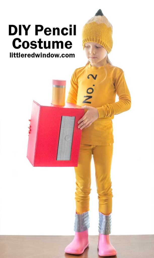 little girl in yellow pencil costume with pencil hat and pink boots holding a red cardboard box pencil sharpener with large pencil in it next to her waist