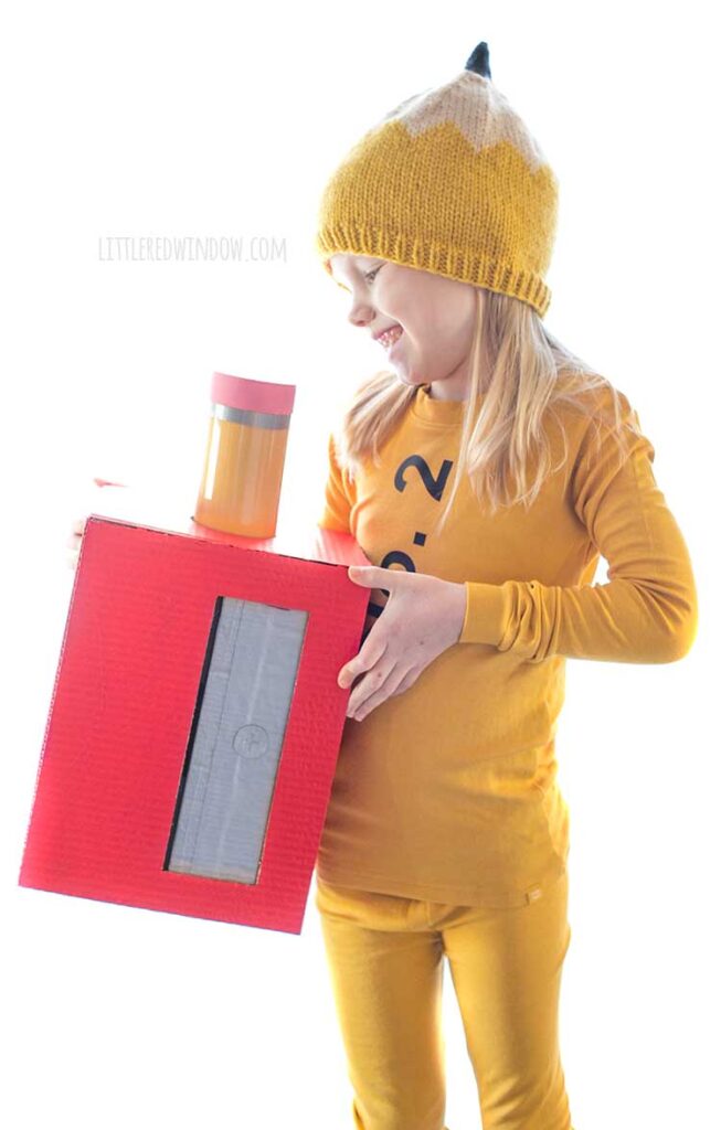 girl in pencil costume looking down and laughing at a large cardboard box pencil sharpener in her hands