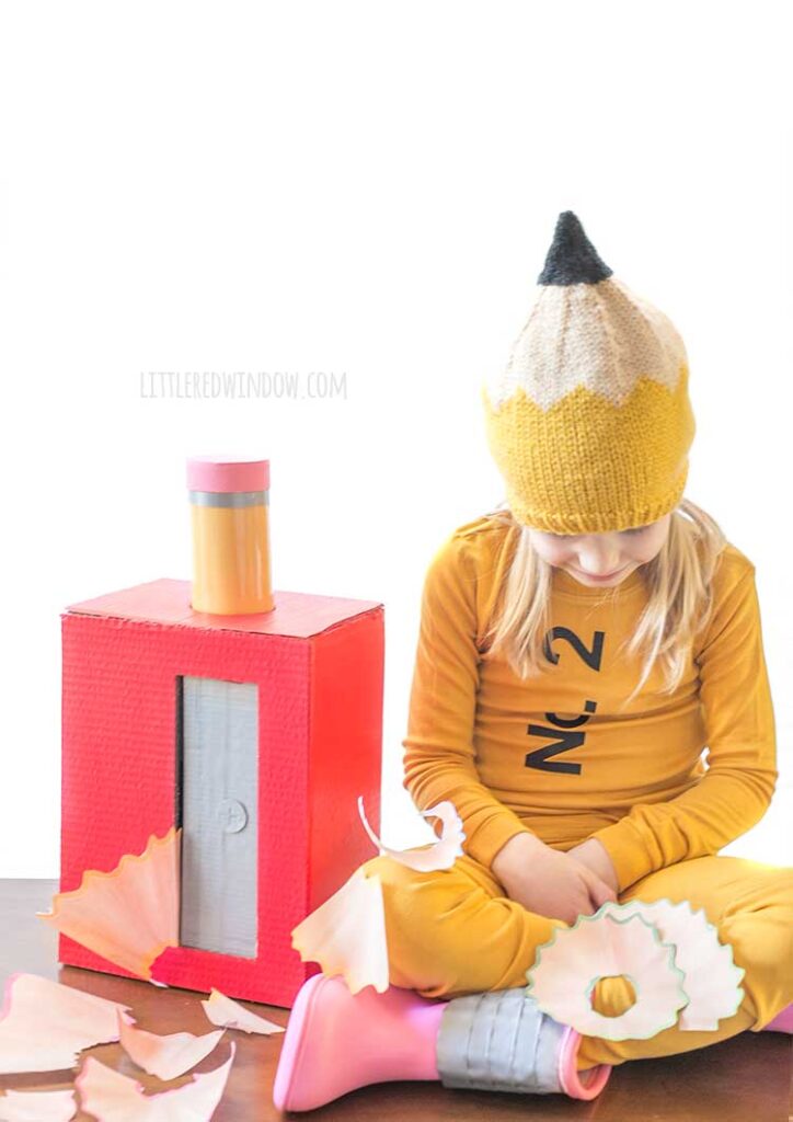 girl sitting with crossed legs wearing a pencil costume on a wood table next to a large red pencil sharpener with a pencil sticking out of the top and large pencil shavings all around her looking down at her lap