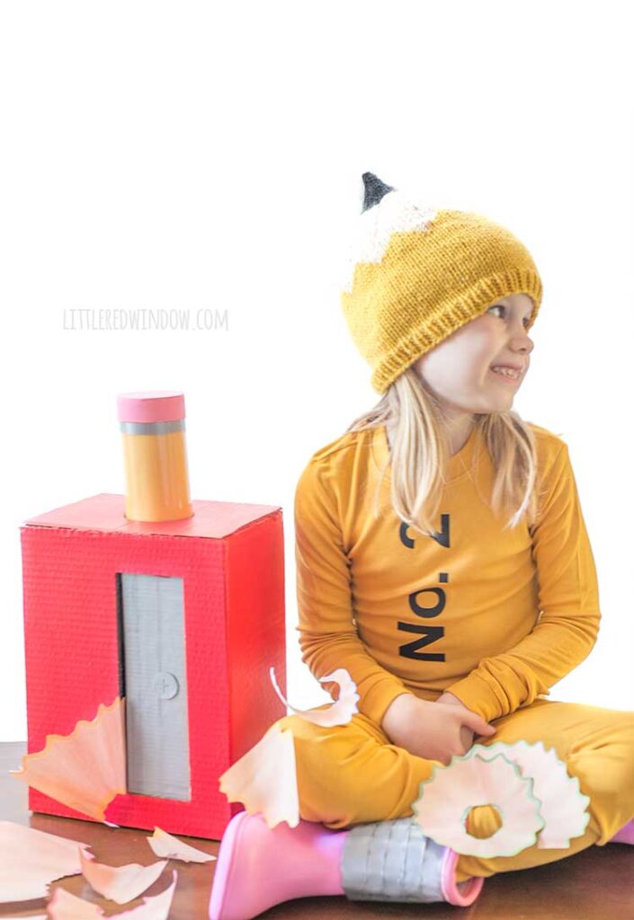 girl sitting with crossed legs wearing a pencil costume on a wood table next to a large red pencil sharpener with a pencil sticking out of the top and large pencil shavings all around her looking off to the right