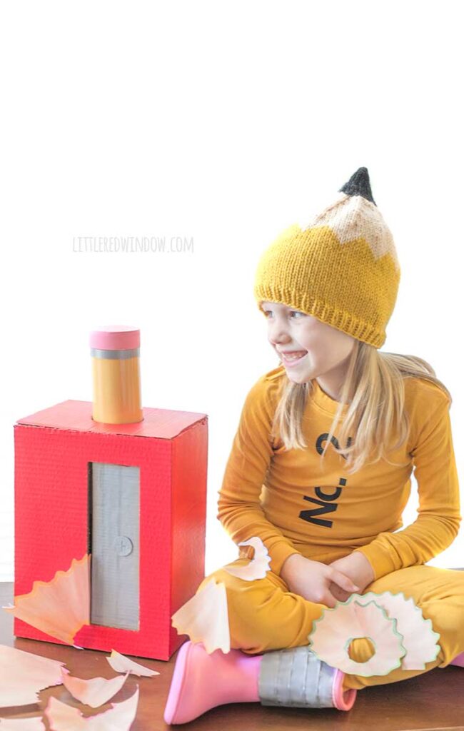 girl sitting with crossed legs wearing a pencil costume on a wood table next to a large red pencil sharpener with a pencil sticking out of the top and large pencil shavings all around her looking off to the left