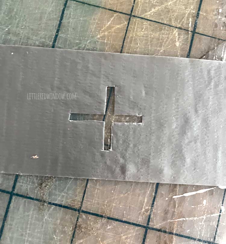 a piece of silver duct tape sitting on a gray cutting mat with a plus sign cut out of the center of the piece