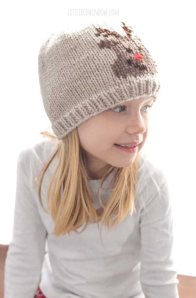 smiling blonde girl in white shirt wearing a tan knit hat with an embroidered reindeer face on the front looking off to the right