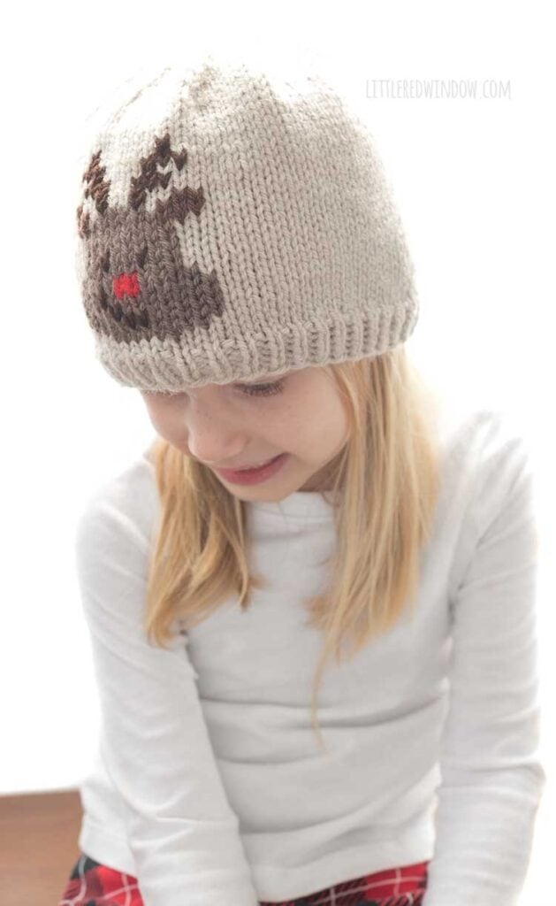 smiling blonde girl in white shirt wearing a tan knit hat with an embroidered reindeer face on the front looking down and to the left