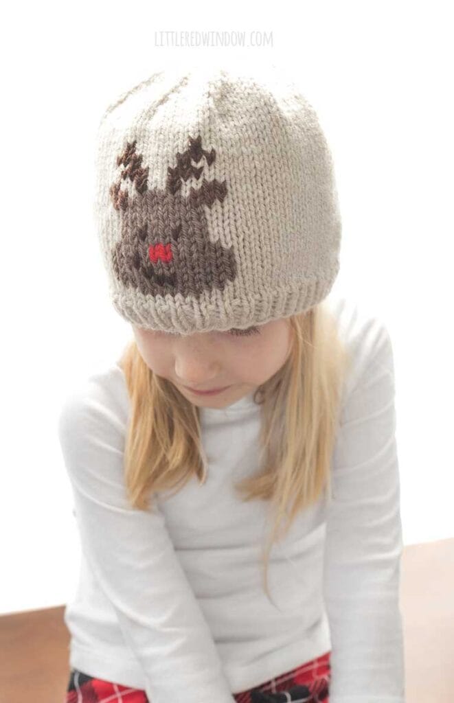smiling blonde girl in white shirt wearing a tan knit hat with an embroidered reindeer face on the front looking down at her lap