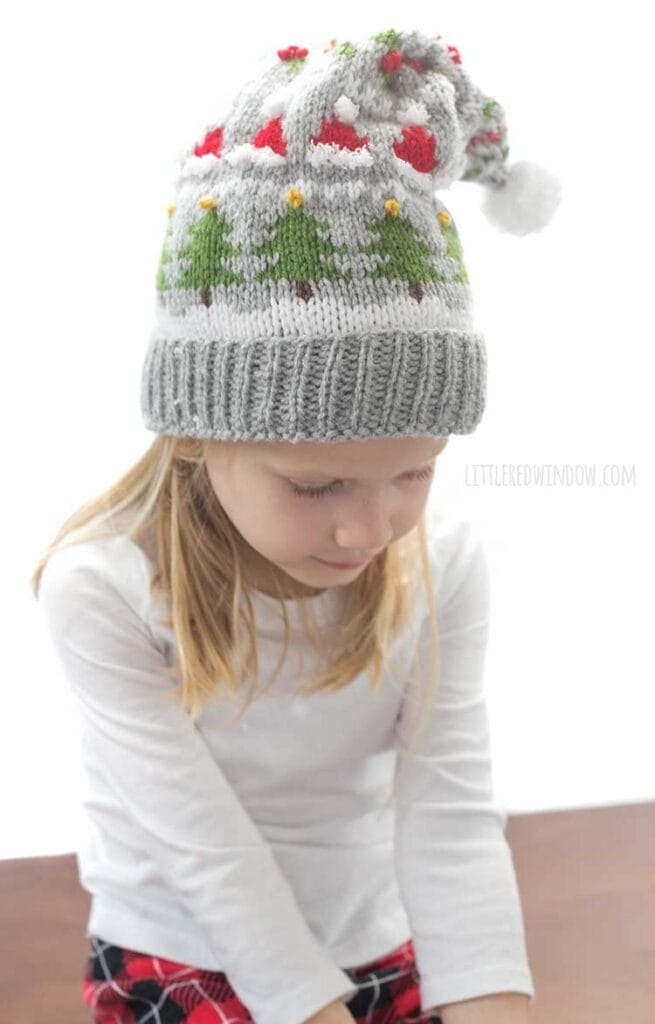 blond girl in white long sleeved shirt wearing a gray knit stocking cap decorated with patterns of Christmas trees Santa hats holly berries and candy cane stripe and a white pom pom on top looking down at her lap