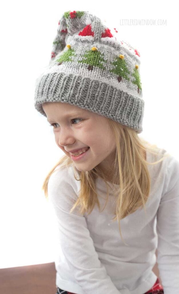 blond girl in white long sleeved shirt wearing a gray knit stocking cap decorated with patterns of Christmas trees Santa hats holly berries and candy cane stripe and a white pom pom on top looking down and off to her left and smiling