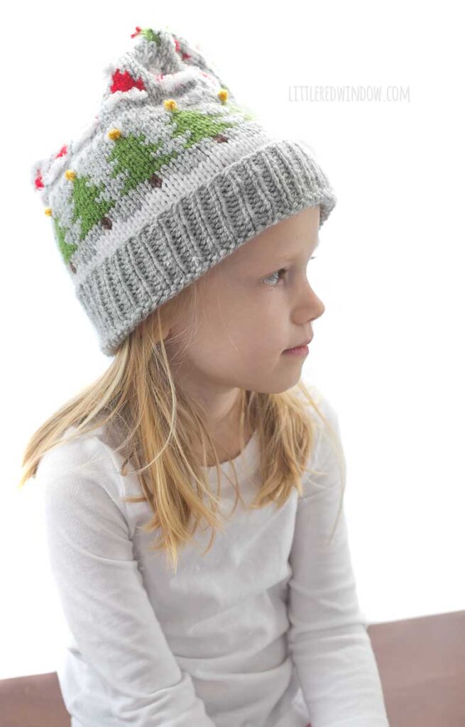 blond girl in white long sleeved shirt wearing a gray knit stocking cap decorated with patterns of Christmas trees Santa hats holly berries and candy cane stripe and a white pom pom on top looking off to the right with stocking cap folded over her shoulder