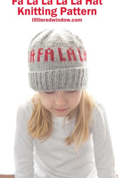 little girl in a white shirt wearing a gray knit hat with folded ribbed brim knit with the words Fa La La La LA around the middle in red looking down at her lap