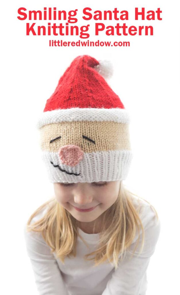 Girl in white shirt wearing a knit hat that looks like Santa's head complete with white beard rosy nose and red stocking cap on top she's looking down and smiling