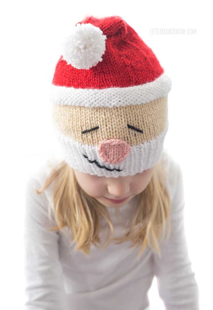 Girl in white shirt wearing a knit hat that looks like Santa's head complete with white beard rosy nose and red stocking cap on top she's looking down