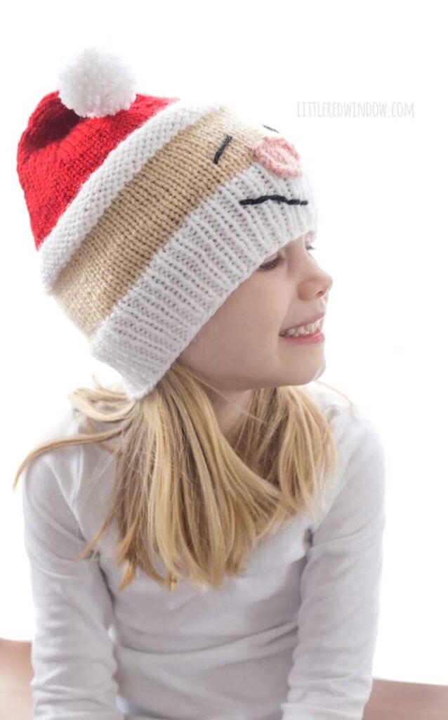 Girl in white shirt wearing a knit hat that looks like Santa's head complete with white beard rosy nose and red stocking cap on top she's looking off to the right and smiling