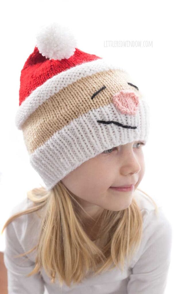 Girl in white shirt wearing a knit hat that looks like Santa's head complete with white beard rosy nose and red stocking cap on top she's looking down and to the right