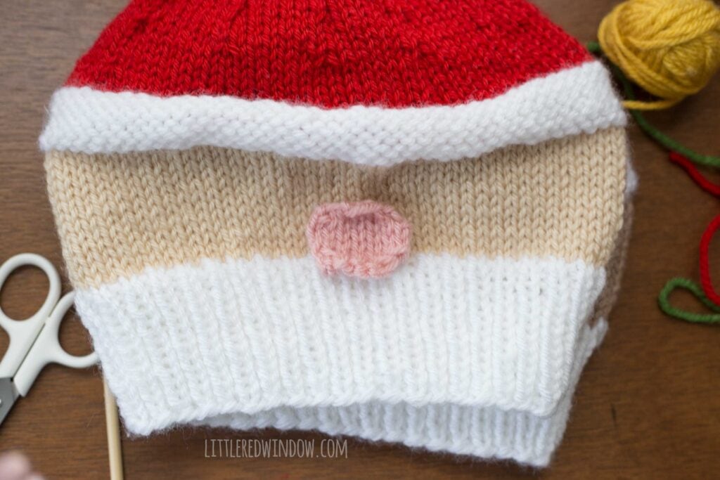 closeup of an in process smiling santa hat showing where to sew the pink nose on to the front of the hat just above the ribbed beard