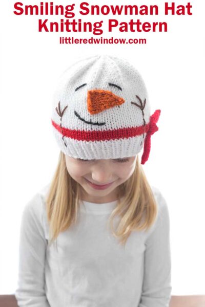 little girl in white shirt with blonde hair wearing a knit hat that looks like a snowman head with carrot nose smile red scarf and stick arms looking down at her lap