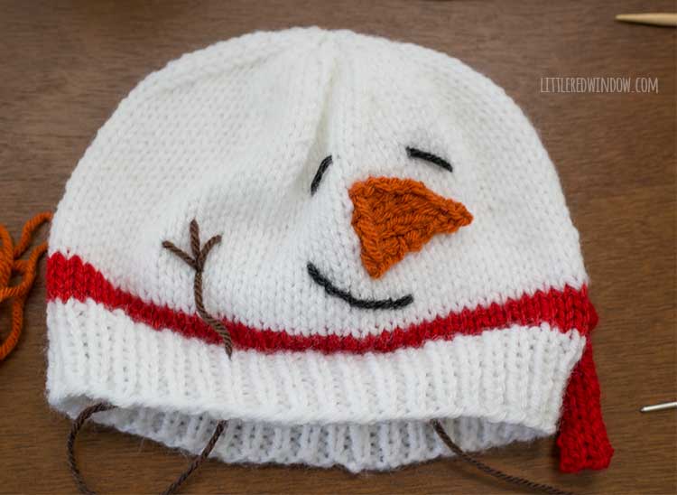snowman hat with brown yarn being used to stitch the stick arms on the side of the face