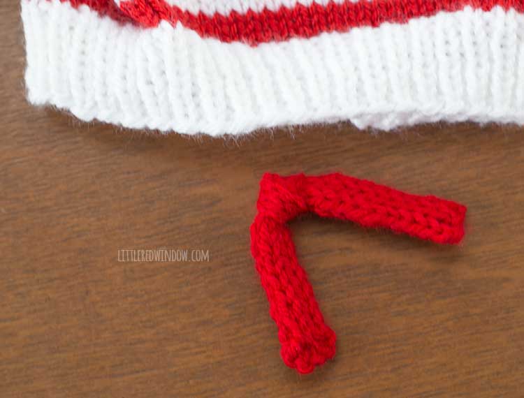 a white knit hat with a thin red strips next to an 8 inch long strip of knitting that is tied in a knot for the smiling snowman hat on a wood surface