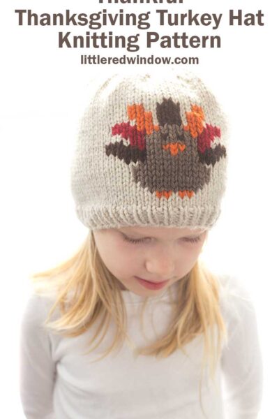 girl in white shirt wearing a tan knit hat with a Thanksgiving Turkey knit on the front