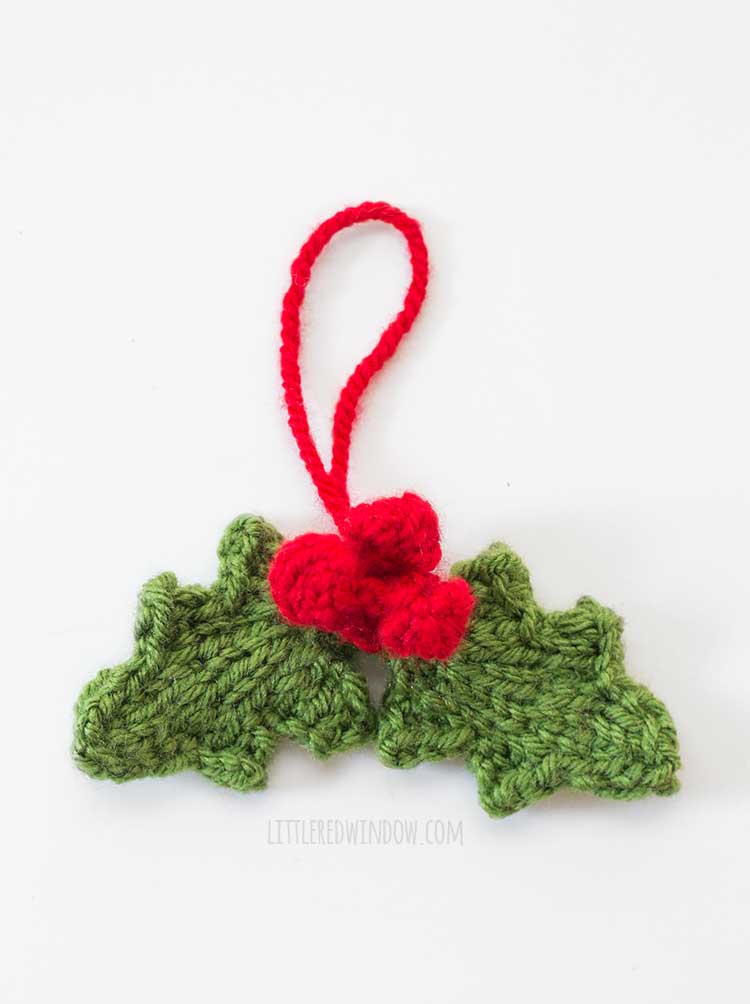 closeup of knit ornament with two green knit holly leaves and three red knit holly berries and a red yarn loop for hanging on a white background