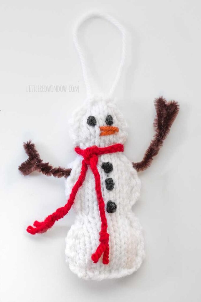 a white knit showman shaped ornament with brown pipe cleaner arms a red yarn braided scarf black eyes and buttons and an orange yarn carrot nose on a white background