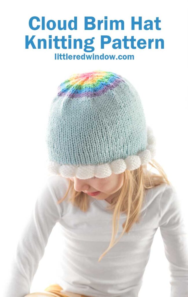 blond girl wearing light blue knit hat with bubble knit white cloud brim and a rainbow decrease section on the top in front of a white background looking down to the left