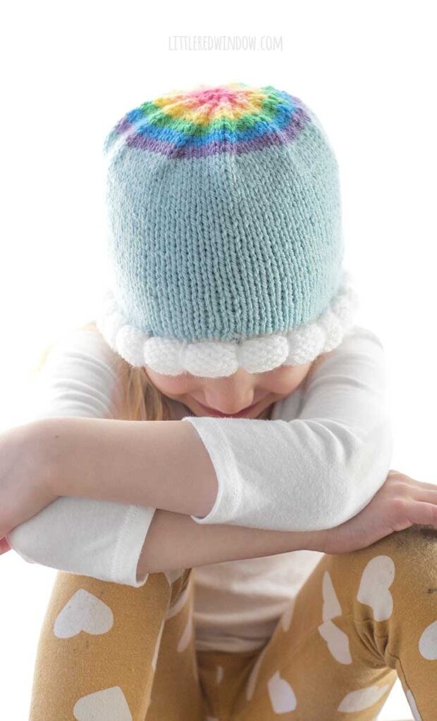 girl with arms crossed and leaning forward wearing the cloud brim knit hat in light blue with a rainbow on top
